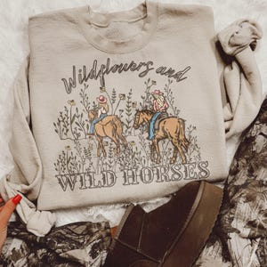 May include: A beige sweatshirt with a graphic print of two women riding horses in a field of wildflowers. The text "Wildflowers and Wild Horses" is printed below the image.