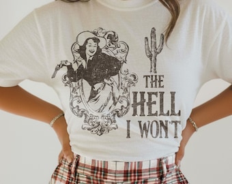 May include: A white short-sleeved t-shirt featuring a black graphic print of a cowgirl in a decorative frame, a cactus, and the text THE HELL I WON'T. The shirt is paired with red, white, and black plaid trousers with a visible button and zip.