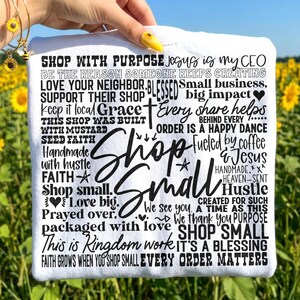 May include: White t-shirt with a black text design that reads "Shop Small" in a large, stylized font. The shirt also features various phrases and words related to faith, small business, and community support.