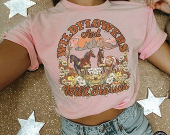 Peut inclure: T-shirt rose avec un imprimé vintage de chevaux sauvages et de fleurs sauvages. Le graphique représente une scène de coucher de soleil avec des montagnes et des cactus. Le texte dit "Wildflowers and Wild Horses".