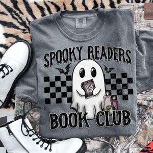 May include: Gray crewneck sweatshirt with the text "SPOOKY READERS BOOK CLUB" and a cartoon ghost holding a book that says "SPELL BOOK". The design includes bats, checkered squares, and candles. White boots are visible.