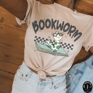 May include: A light beige t-shirt with the text "BOOKWORM" in black lettering. A cartoon green worm wearing glasses is on top of a book with a checkered pattern.