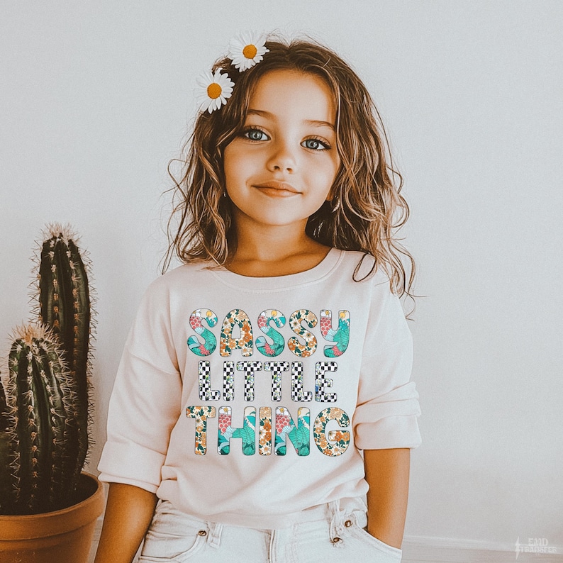 Puede incluir: Una ni&ntilde;a peque&ntilde;a con una camiseta rosa claro de manga larga con las palabras "SASSY LITTLE THING" en un dise&ntilde;o floral y a cuadros. Tiene flores de margarita en el pelo. Se ve un cactus.