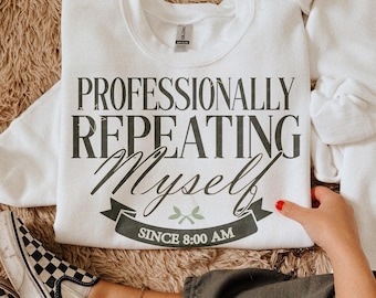May include: A white crewneck sweatshirt featuring dark gray text that reads PROFESSIONALLY REPEATING Myself and a banner graphic with the text SINCE 8:00 AM. The design includes a small green leaf icon centered above the banner on the front.
