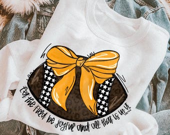 May include: A white crewneck sweatshirt featuring a graphic of a brown leopard print football adorned with a large yellow bow. The football has black and white star-patterned stripes. The text reads, Let the field be joyful and all that is in it.