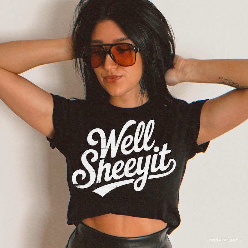 Puede incluir: Top corto negro con el texto "Well Sheeeyit" en letras blancas estilizadas. La camiseta es una prenda informal. La modelo lleva gafas de sol con cristales naranjas.
