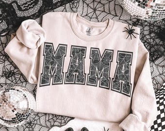 May include: A light beige crewneck sweatshirt featuring the word MAMA in large, block-style letters. The letters are filled with a black and white spiderweb pattern and outlined in black. The garment has long sleeves and a ribbed collar.