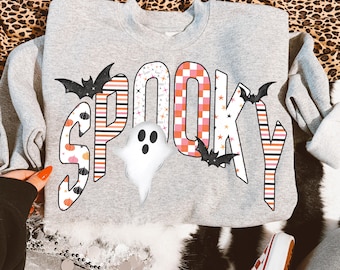 May include: A heather grey crewneck sweatshirt featuring the word SPOOKY in large, decorative letters. The letters are filled with patterns of black and orange stripes, pumpkins, stars, and a pink and orange checkerboard. A white ghost and black bats are included.