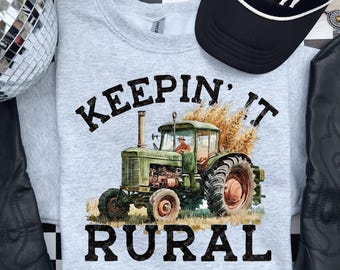 May include: A light gray sweatshirt with the words "KEEPING IT RURAL" in black block letters. The sweatshirt features a green tractor graphic with a farmer driving it. A black baseball cap is visible in the background.