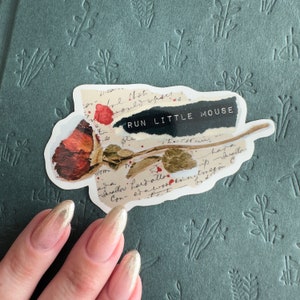 May include: A sticker with a dried rose and a black and white paper background. The text "RUN LITTLE MOUSE" is printed on the sticker.
