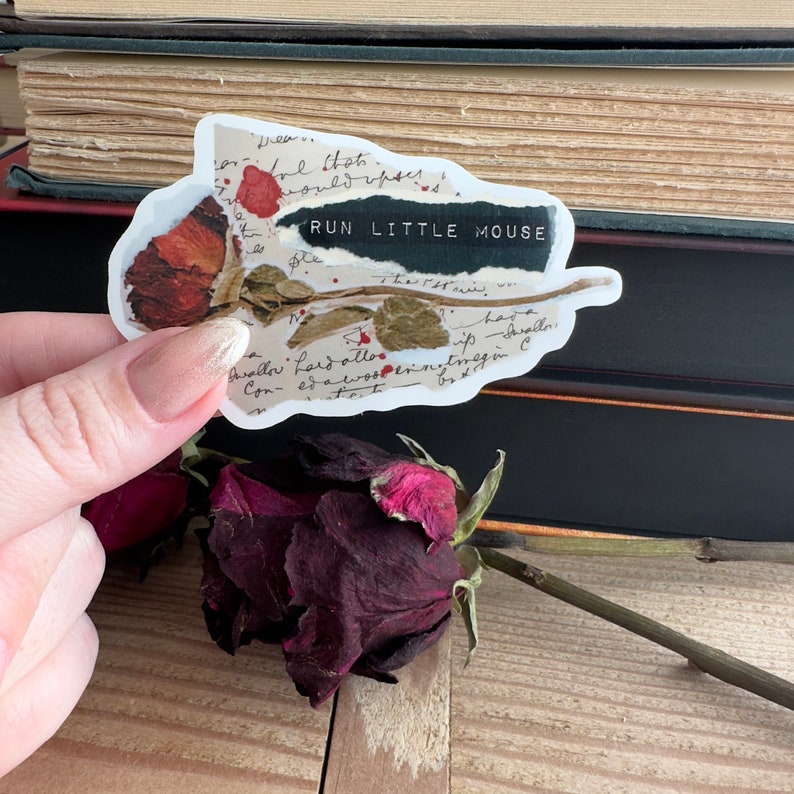 May include: A white sticker with a dried rose and handwritten text that reads "RUN LITTLE MOUSE". The sticker is placed on a stack of books.