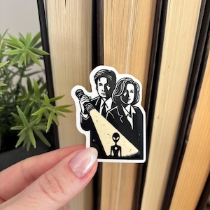May include: Black and white sticker featuring a stylized illustration of two people holding a flashlight, shining it on a small alien figure. The sticker is on a book.