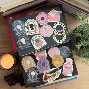 May include: A collection of colorful stickers featuring various designs, including a book club logo, a cherry design, a book with the text "Good Girls Book Club", a sticker with the text "Finer Things", a sticker with the text "Shadow Daddy", a sticker with the text "Violent Little Thing", and a sticker with the text "I Will Not Die Today".
