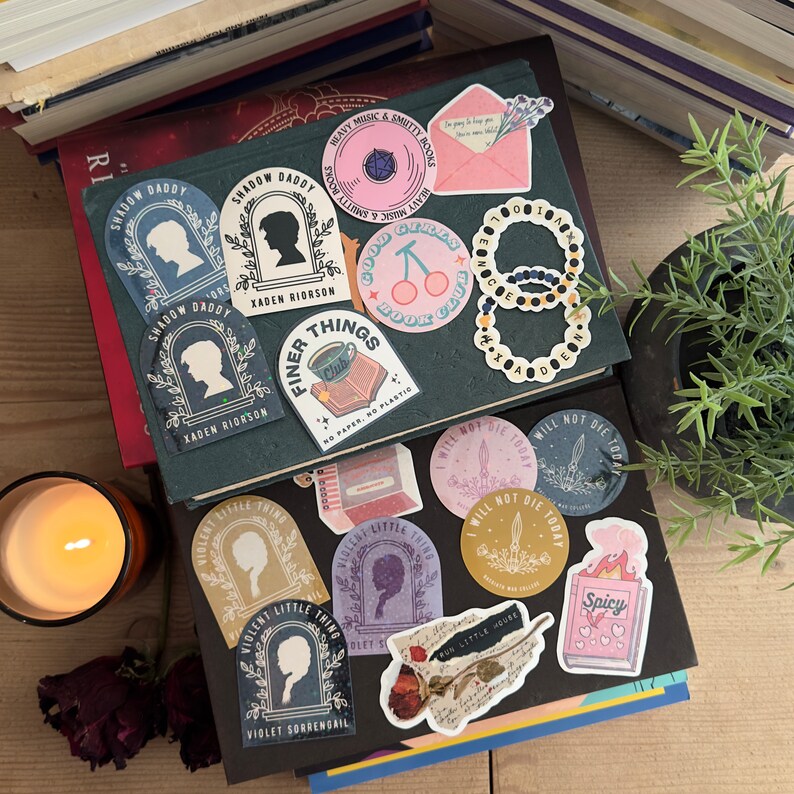 May include: A collection of colorful stickers featuring various designs, including a book club logo, a cherry design, a book with the text "Good Girls Book Club", a sticker with the text "Finer Things", a sticker with the text "Shadow Daddy", a sticker with the text "Violent Little Thing", and a sticker with the text "I Will Not Die Today".