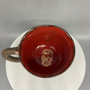 Könnte beinhalten: Eine handgefertigte Keramik-Tasse mit roter Innenseite und brauner Außenseite. Die Tasse hat ein goldenes Totenkopf-Design in der Mitte und einen braunen Henkel. Die Tasse steht auf einem weißen Teller.