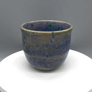 May include: A handcrafted ceramic cup with a speckled blue and grey glaze. The cup has a rounded form and a slightly flared rim. Green and yellow speckles add visual interest to the surface.