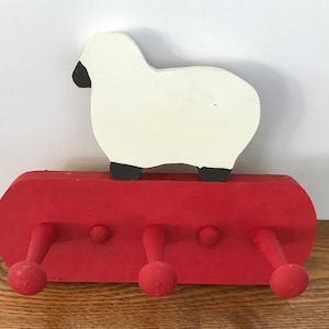 May include: A red wooden coat rack with four hooks and a white wooden sheep on top. The sheep has black eyes and hooves.
