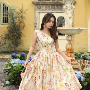 May include: A sleeveless, knee-length dress with a floral pattern of pink, yellow, and green tulips on a cream background. The dress has a fitted bodice and a flared skirt. The woman is wearing gold sandals.