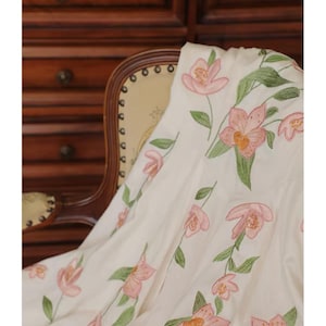 May include: White fabric featuring embroidered pink flowers and green leaves. The floral design is arranged vertically. The word "FABRIC" is at the top of the image. The fabric is draped over a chair.