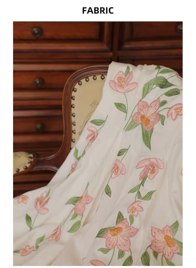 May include: White fabric featuring embroidered pink flowers and green leaves. The floral design is arranged vertically. The word "FABRIC" is at the top of the image. The fabric is draped over a chair.