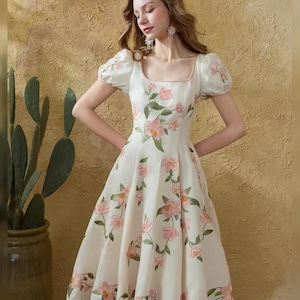May include: A white dress featuring a square neckline and short, puffed sleeves. The dress is decorated with pink floral embroidery and green leaves. The dress has a fitted bodice and a flared skirt, creating a classic silhouette.