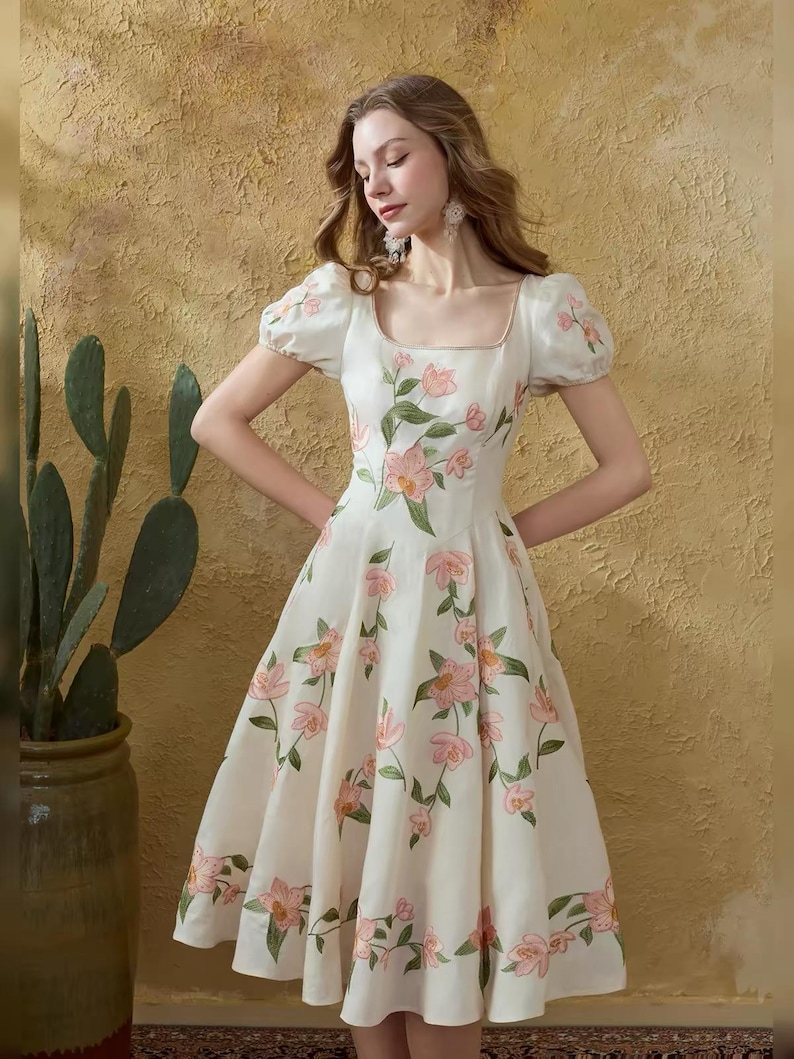 May include: A white dress featuring a square neckline and short, puffed sleeves. The dress is decorated with pink floral embroidery and green leaves. The dress has a fitted bodice and a flared skirt, creating a classic silhouette.
