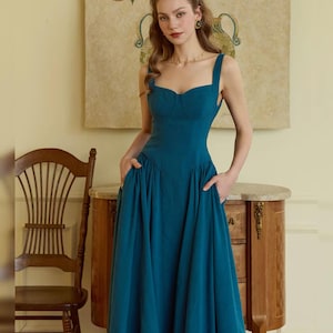 May include: A teal-colored midi dress with a sweetheart neckline and wide straps. The dress features a fitted bodice and a flared skirt with pockets. The model is standing in front of a wooden cabinet and a painting.