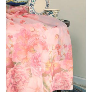 May include: A sheer pink floral tablecloth with large peony blooms in shades of pink and peach. The tablecloth drapes over a table, with a glimpse of a blue patterned rug and decorative china in the background.