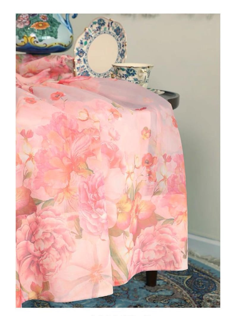 May include: A sheer pink floral tablecloth with large peony blooms in shades of pink and peach. The tablecloth drapes over a table, with a glimpse of a blue patterned rug and decorative china in the background.