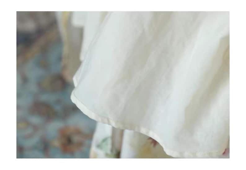 May include: Close-up of a flowing, off-white fabric with a soft, translucent quality. The fabric has a slightly ruffled edge and appears to be part of a garment. The background is blurred, suggesting a focus on the textile's texture and drape.