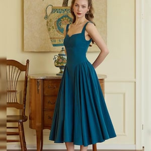 May include: A teal-colored midi dress with a fitted bodice and a flared skirt. The dress features a sweetheart neckline and wide straps. The model is wearing gold sandals. The background includes a wooden chair, a decorative table, and a painting.