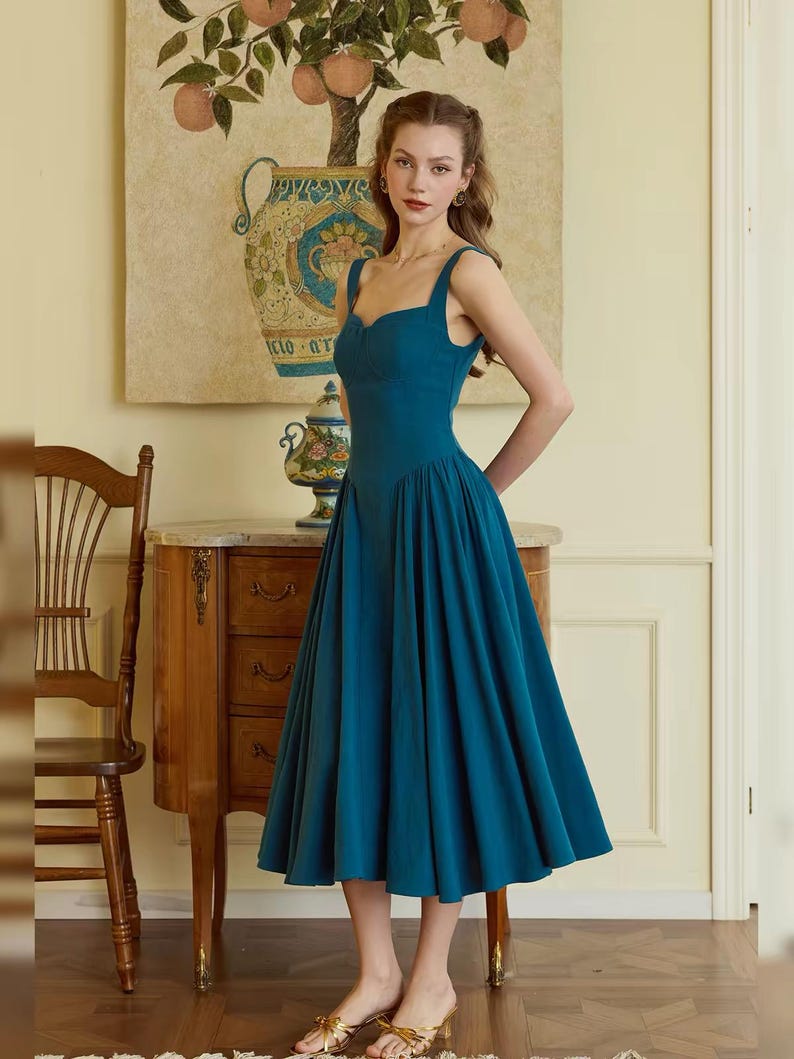 May include: A teal-colored midi dress with a fitted bodice and a flared skirt. The dress features a sweetheart neckline and wide straps. The model is wearing gold sandals. The background includes a wooden chair, a decorative table, and a painting.