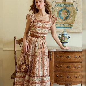 May include: A cream-colored midi dress with a brown floral pattern and a brown belt. The dress has a square neckline, ruffled sleeves, and a tiered skirt. A small, woven handbag hangs from the shoulder. The dress is worn by a person.