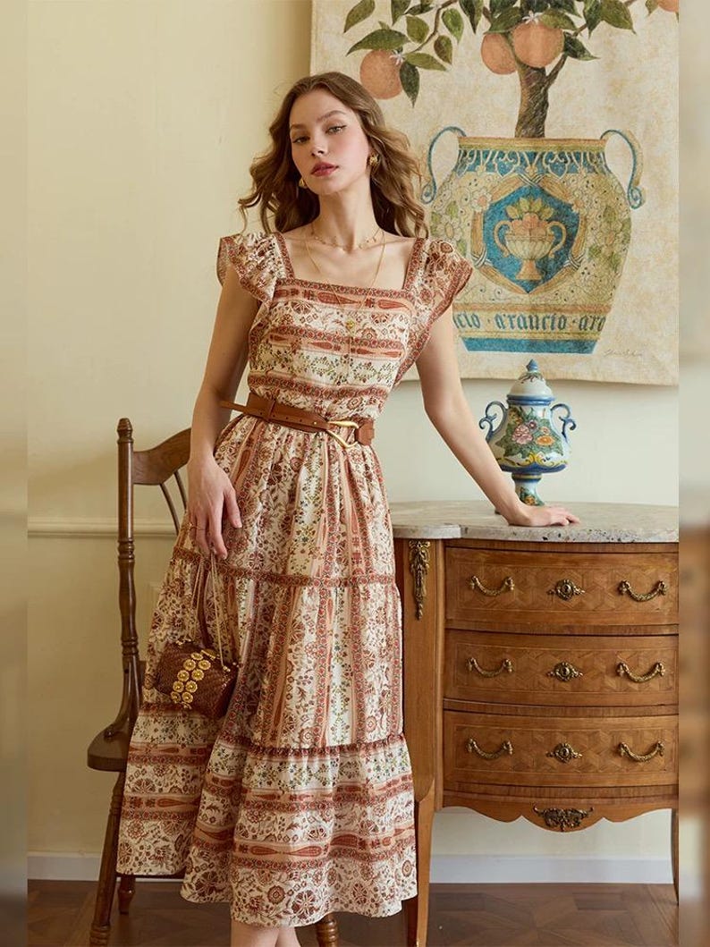 May include: A cream-colored midi dress with a brown floral pattern and a brown belt. The dress has a square neckline, ruffled sleeves, and a tiered skirt. A small, woven handbag hangs from the shoulder. The dress is worn by a person.
