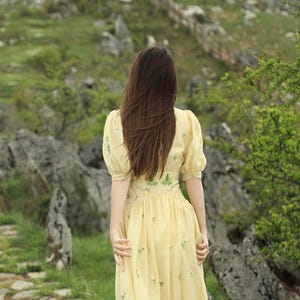 Puede incluir: Un vestido amarillo claro con mangas cortas abullonadas y bordados florales. La persona lleva el vestido y camina por un sendero de piedra. El fondo presenta rocas, vegetaci&oacute;n y una valla de madera.