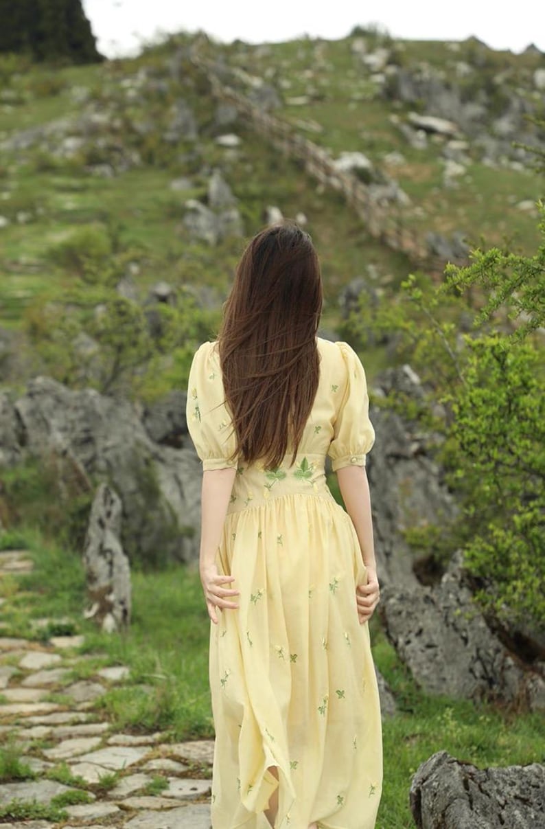 Puede incluir: Un vestido amarillo claro con mangas cortas abullonadas y bordados florales. La persona lleva el vestido y camina por un sendero de piedra. El fondo presenta rocas, vegetaci&oacute;n y una valla de madera.
