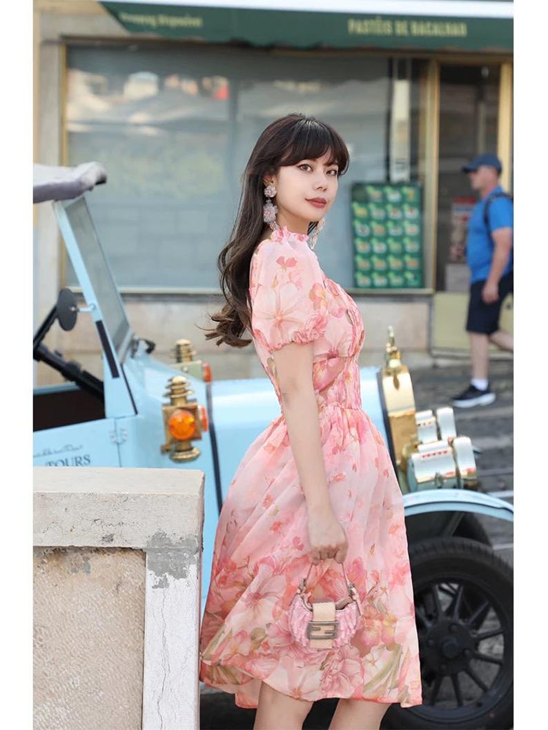 May include: A pink floral print dress with short sleeves and a cinched waist. The dress is paired with a small, pink floral handbag. A vintage light blue car is in the background.