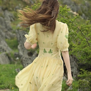 Puede incluir: Un vestido amarillo claro con mangas cortas y una falda fluida. El vestido presenta bordados florales verdes y una cintura ajustada. La persona camina en un entorno natural al aire libre con rocas y vegetaci&oacute;n.