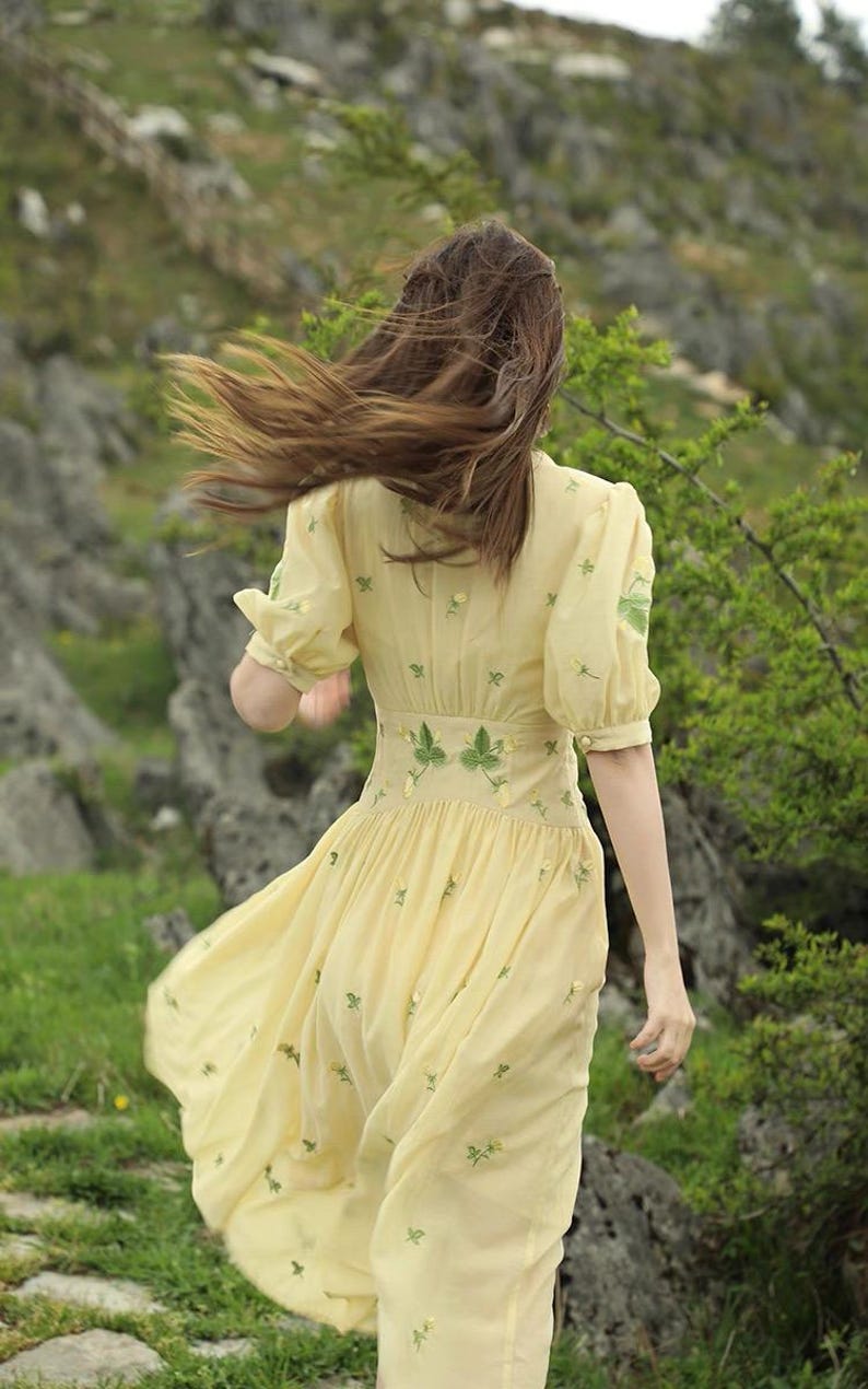 Puede incluir: Un vestido amarillo claro con mangas cortas y una falda fluida. El vestido presenta bordados florales verdes y una cintura ajustada. La persona camina en un entorno natural al aire libre con rocas y vegetaci&oacute;n.