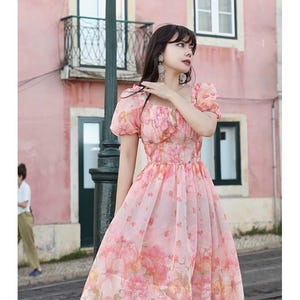 May include: A pink floral print dress with short, puffy sleeves and a square neckline. The dress features a fitted bodice and a flowing skirt, with a knee-length hemline. The background includes a pink building and a street.