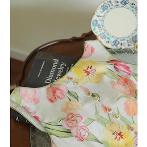 May include: A white top adorned with a watercolour floral design of pink and yellow tulips. The top features small buttons and is displayed on a dark wooden surface. A book titled "Diamond Jewelry" is visible.