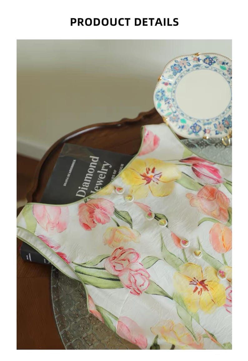 May include: A white top adorned with a watercolour floral design of pink and yellow tulips. The top features small buttons and is displayed on a dark wooden surface. A book titled "Diamond Jewelry" is visible.