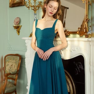 May include: A teal-colored formal dress with a sweetheart neckline and wide straps. The dress has a fitted bodice and a flowing skirt. The woman is wearing gold jewelry. The background includes a fireplace, a mirror, and a decorative chair.