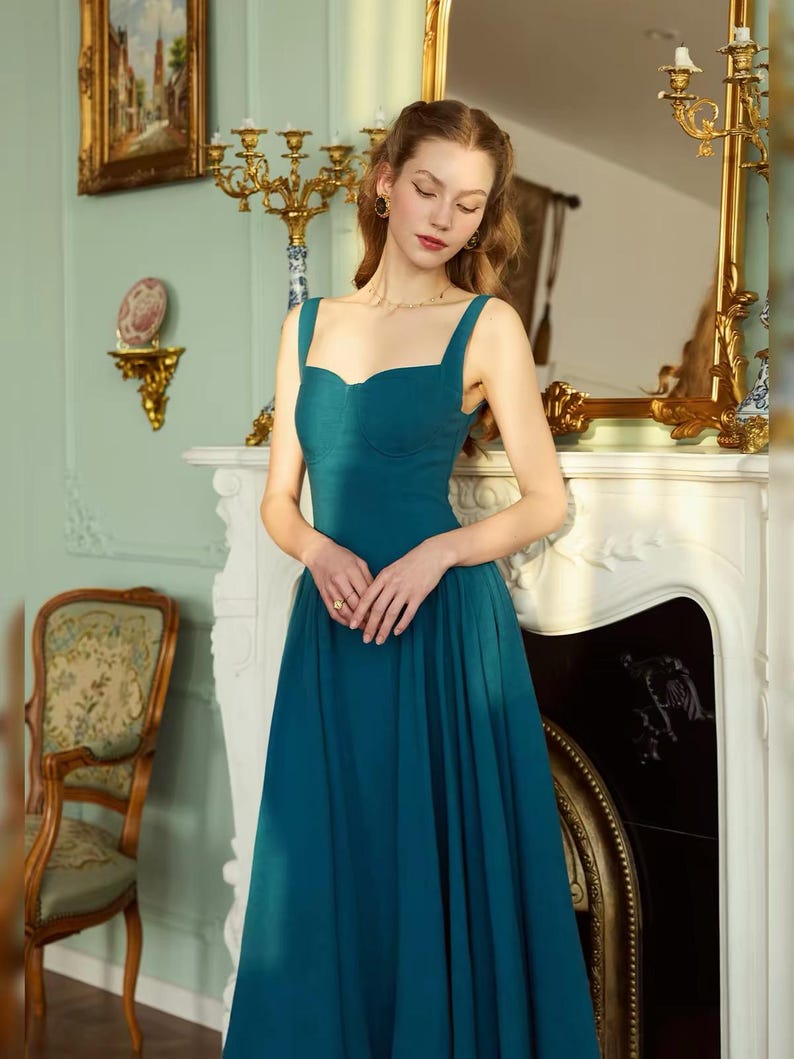 May include: A teal-colored formal dress with a sweetheart neckline and wide straps. The dress has a fitted bodice and a flowing skirt. The woman is wearing gold jewelry. The background includes a fireplace, a mirror, and a decorative chair.