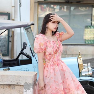 May include: A pink floral print dress with short puff sleeves and a square neckline. The dress is knee-length and has a full skirt. A small pink handbag is held in the hand. A light blue vintage car is in the background.