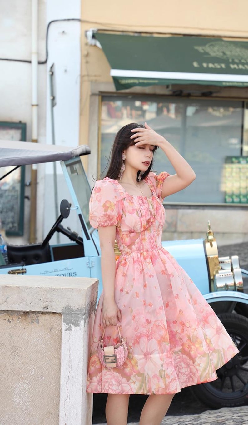 May include: A pink floral print dress with short puff sleeves and a square neckline. The dress is knee-length and has a full skirt. A small pink handbag is held in the hand. A light blue vintage car is in the background.