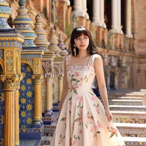 May include: A cream-coloured, sleeveless dress with a floral pattern of pink roses and green leaves. The dress has a square neckline and a full skirt. The background features ornate architecture with columns and decorative tiles.