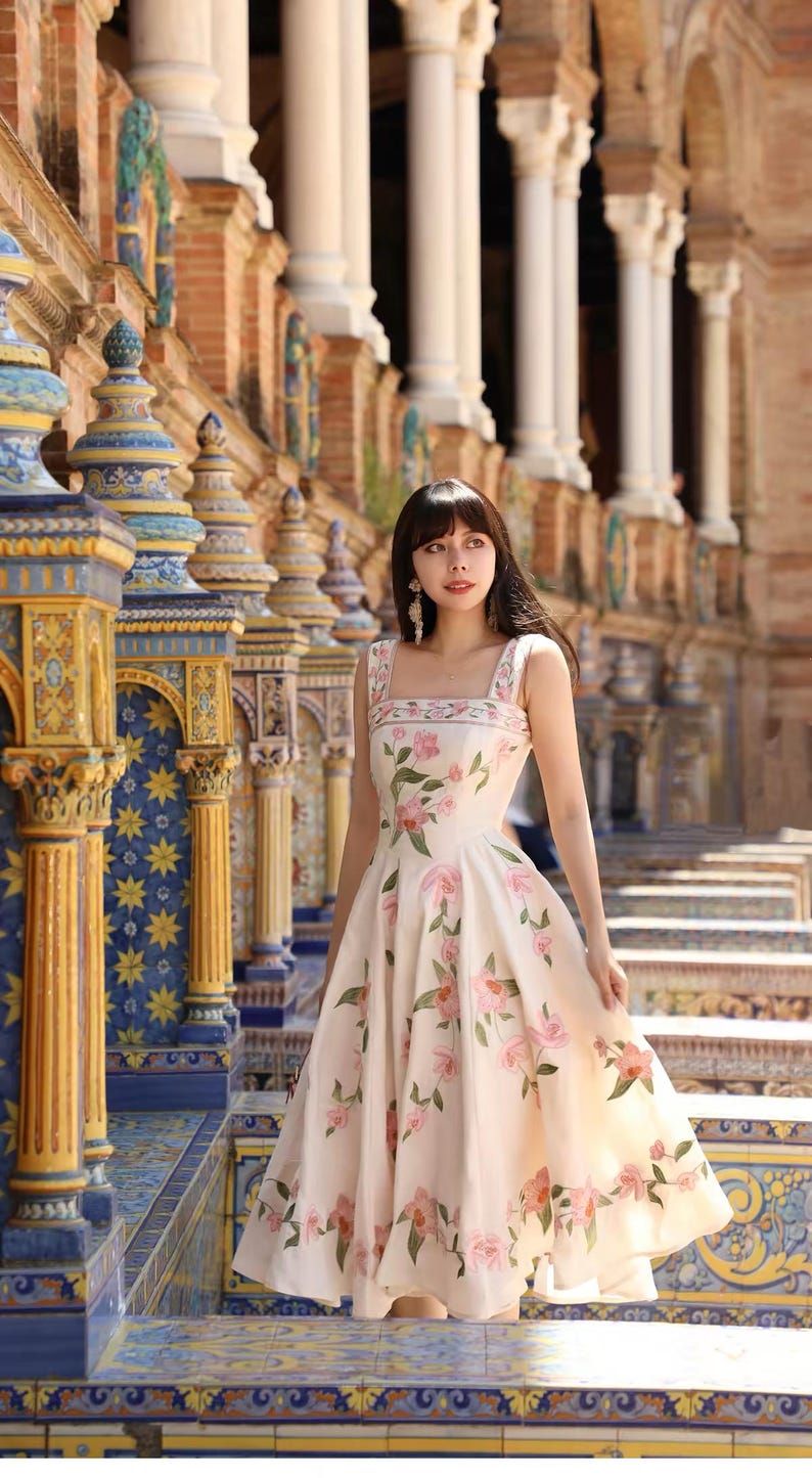 May include: A cream-coloured, sleeveless dress with a floral pattern of pink roses and green leaves. The dress has a square neckline and a full skirt. The background features ornate architecture with columns and decorative tiles.