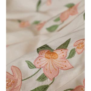 May include: Close-up of off-white fabric with embroidered pink flowers and green leaves. The floral embroidery is detailed, with a focus on the petals and the texture of the thread. The fabric is slightly creased.