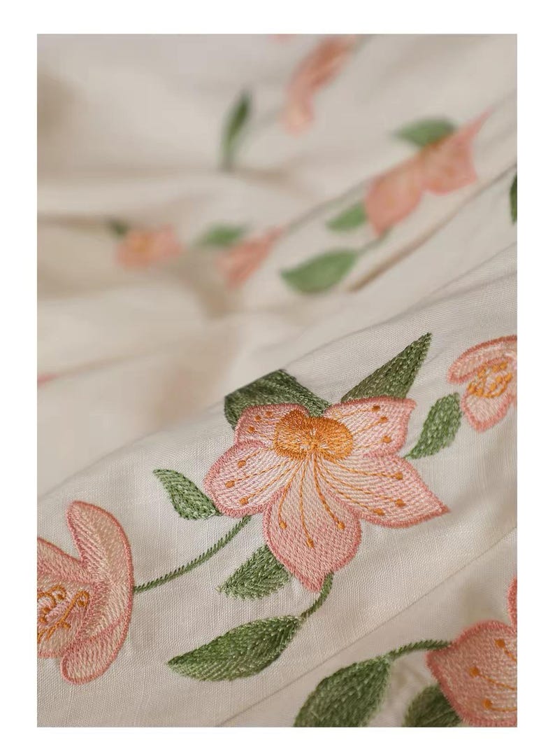 May include: Close-up of off-white fabric with embroidered pink flowers and green leaves. The floral embroidery is detailed, with a focus on the petals and the texture of the thread. The fabric is slightly creased.